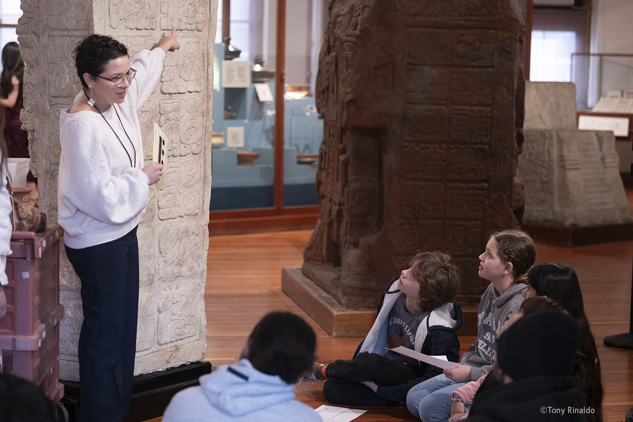 educator points to glyph on large maya sculpture in front of students in gallery.