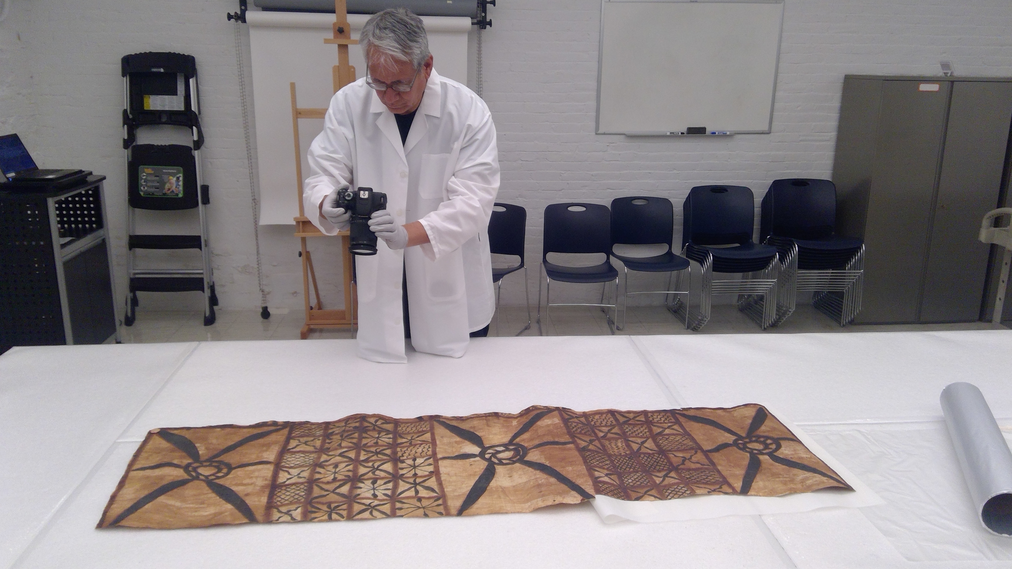 Dan Taulapapa wears a white lab coat and takes a photo of a siapo (bark tapa cloth).