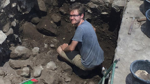harvard student archaeologist smiles from deep excavation.