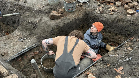 2 archaeologists digging in deep pit.