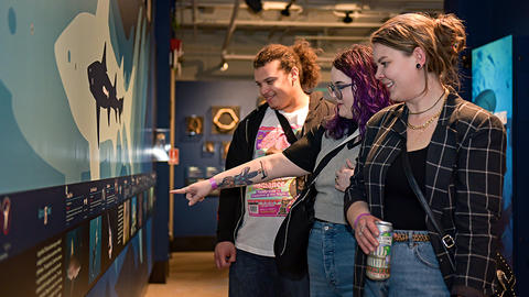 3 young adults smile and gesture at gallery display.