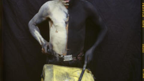 A child plays a yellow drum. Standing against a black background, their body is painted black and white, splitting their body vertically down the middle.