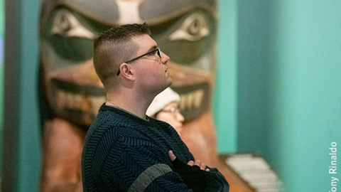 profile of man and person looking at gallery with memorial pole int the background.