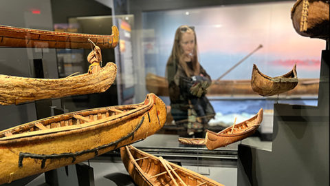 woman looks at canoe models with a large canoe in background.