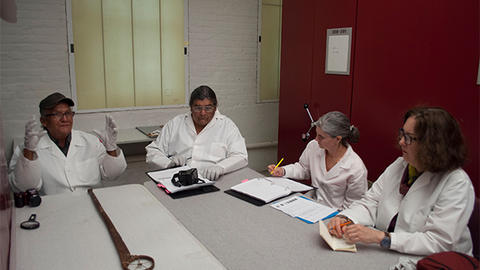 Four people sit around a table in white lab coats surrounding a long staff laid flat on the table. A person all the way to the left holds their hands up as if telling a story. Others take notes.