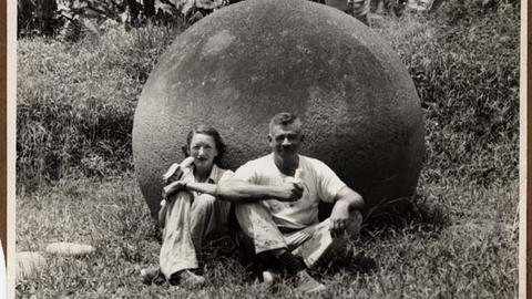 Two people sit in front of large stone sphere eating bananas.