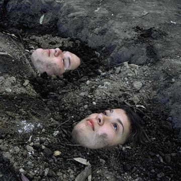 Two girls lie buried face up in the dirt, only their faces are visible. One girl lays with her eyes open, while the other's are shut.