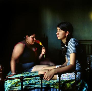 Two women sit in a dark room on a bed with a bright green leaf-patterned comforter.
