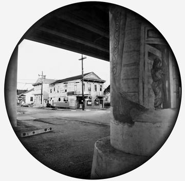 A black and white photo of a house from behind a column across the street. A mural is painted on the column of a woman in a doorway holding a child. The photo is cropped into a perfect circle, with black edges that fade into the photo.