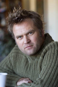 Stephen Dupont looks into the camera lens. His hair is spiked up in the front and he wears an olive green sweater.