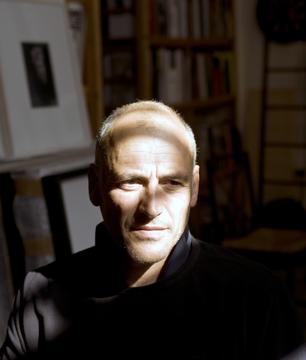 Guy Tillim looks to the right of the camera. Light comes in from a window with blinds, creating stripes of shadow across his face.