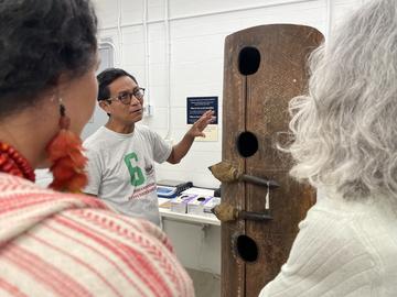 over the shoulders of two people, a man gestures at a large cylindrical drum