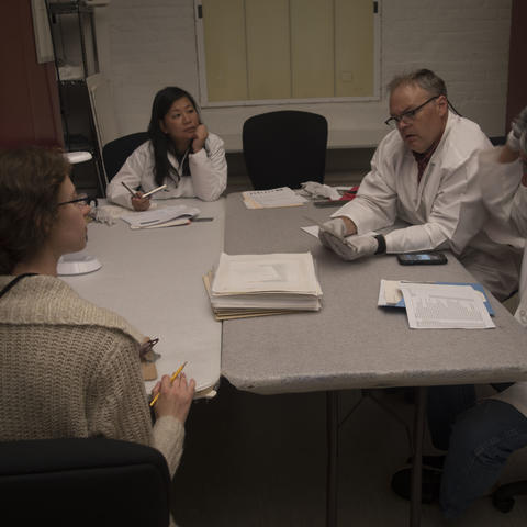 Four people sit around a table, three wearing lab coats. One person on the far right raises their hands to point at their head, as if explaining the use of an object.