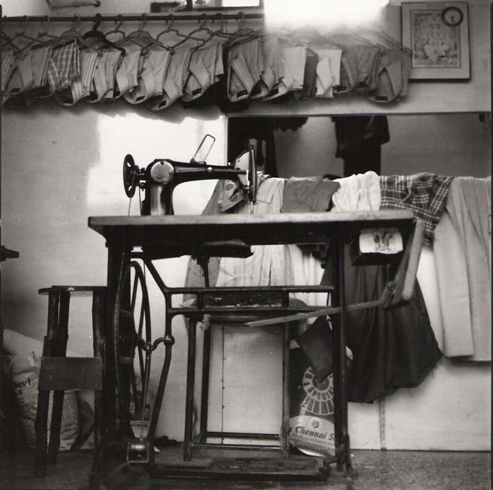 Black and white photo of a sewing machine table taken from a low angle. The wall has several rows of neatly folded clothes hanging from it.