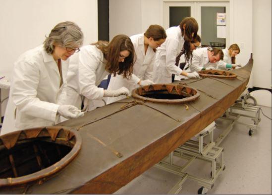 A large brown kayak cuts diagonally across the picture. Researchers in white lab coats peer over its surface, analyzing its bow and openings for passengers.
