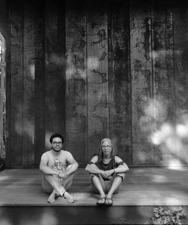 Ilana and Rodrigo of TRES Art sit with their knees up on a raised wooden floor. They are pictured in black and white.