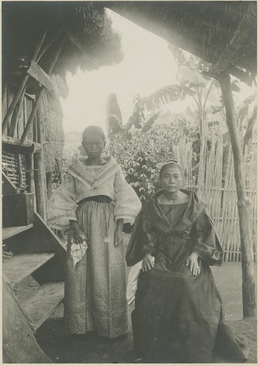 girl stands next to seated woman both looking at lens in front of village structures.