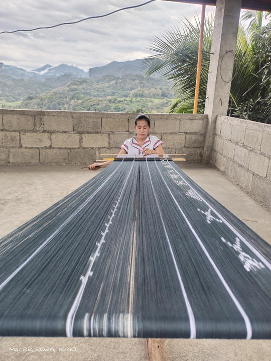 woman seated on porch with verdant mountains in background weaves a long blanket.