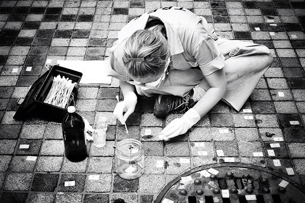 In black and white, a woman leans over a tiled floor wearing glasses. She holds a long Q-tip and is surrounded by tiles she seems to have labelled.