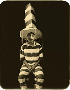In a sepia-toned photo, a black woman sits with hands folded in her lap facing the camera. Wearing a striped jumpsuit. She wears a tall hat with matching stripes, with the left half of her face painted white.