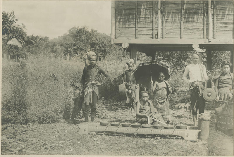 filipinos with instruments, two hold a dangling gong, one kneels behind six gongs in a flat holder, others hold umbrellas.
