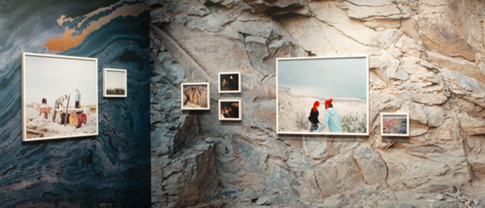 Two walls of an exhibit join at the center. One wall is painted in an abstract ocean, the other appears as a rock face. There are frames photos of varying sizes in a straight line along both walls.