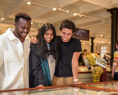 three smiling college students look at gallery case together with more people behind.