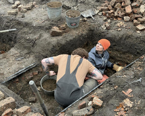 2 archaeologists digging in deep pit.
