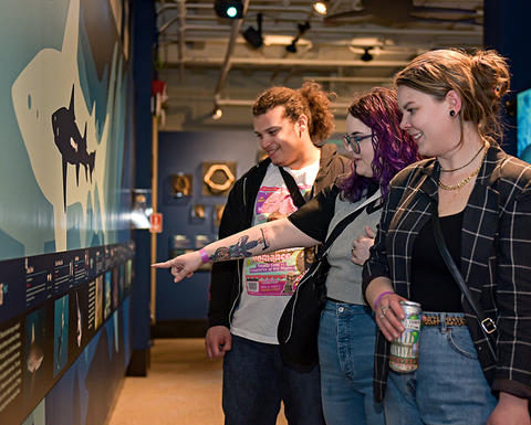3 young adults smile and gesture at gallery display.