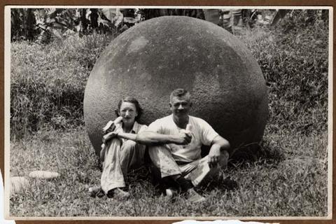 Two people sit in front of large stone sphere eating bananas.